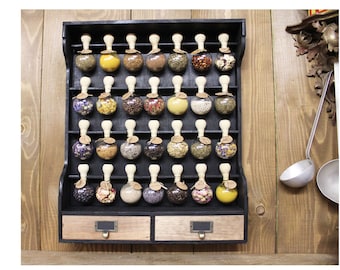 May include: A black wooden spice rack with two drawers, holding 36 small glass jars with various spices and herbs. The jars have wooden lids with a knob on top. The rack is mounted on a wooden wall.