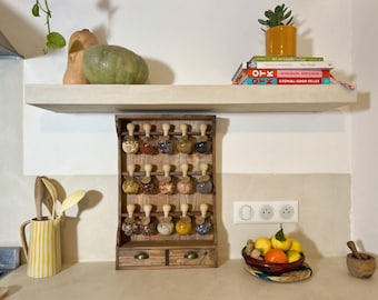 Puede incluir: Un especiero de madera oscura con tres barras horizontales que sostienen quince frascos de vidrio con tapones de madera clara. El mueble cuenta con dos pequeños cajones en la base con tiradores de metal. Cada frasco contiene especias.