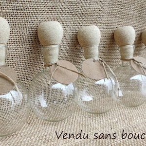 May include: Six clear glass bottles with cork stoppers and brown paper labels. The bottles are arranged in a row on a brown hessian background. The text "Vendu sans bouchons!" is visible at the bottom of the image.