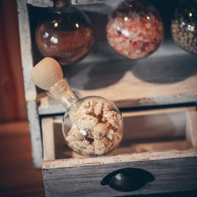 May include: A white wooden shelf with a drawer open, revealing a glass bottle filled with dried white flowers. The bottle has a wooden stopper and is sitting on a white surface. There are three other glass bottles on the shelf, filled with various items, including red and pink crystals.