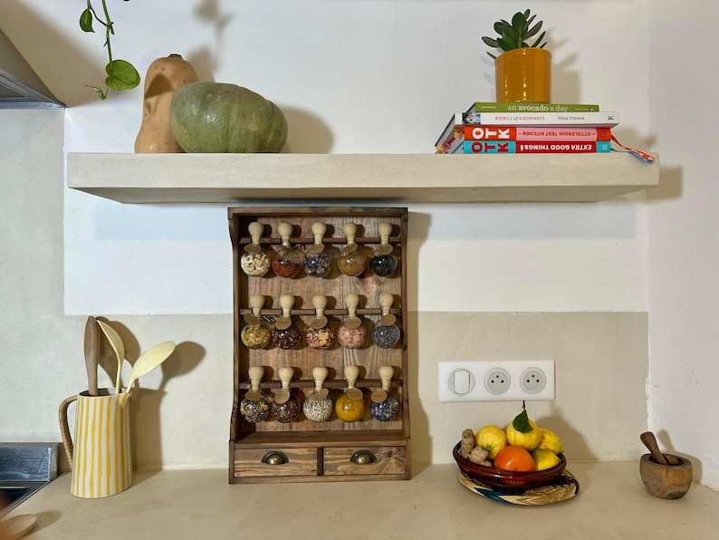 Puede incluir: Un estante de especias de madera con 18 tarros de vidrio llenos de diferentes especias. El estante tiene dos cajones y est&aacute; montado en una pared blanca. Hay dos calabazas amarillas y una suculenta en maceta en un estante blanco sobre el estante.