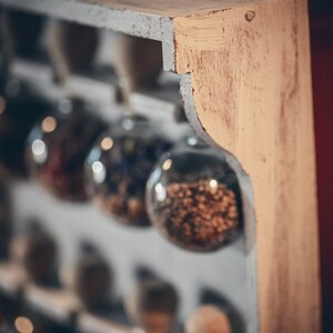 May include: A wooden shelf with glass jars filled with various dried herbs and spices. The jars are hanging from the shelf and are arranged in a row. The shelf is painted white with a light brown stain on the edge.