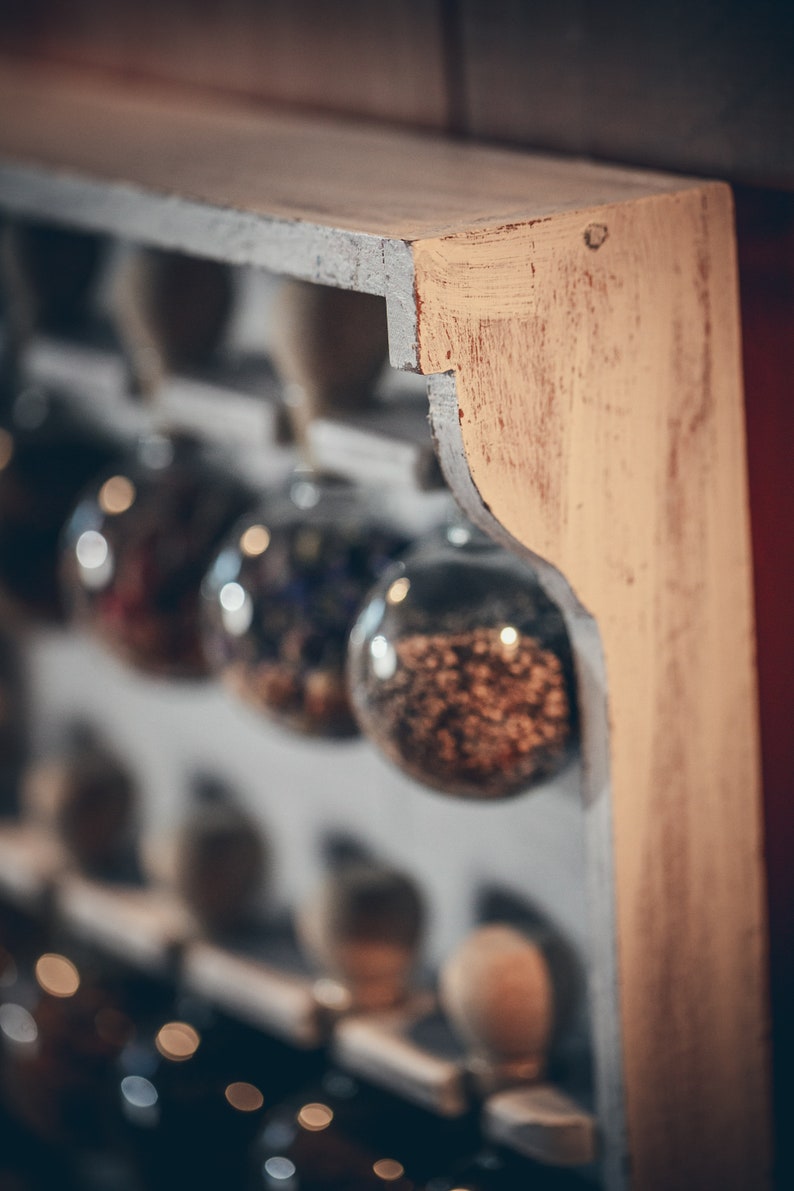 May include: A wooden shelf with glass jars filled with various dried herbs and spices. The jars are hanging from the shelf and are arranged in a row. The shelf is painted white with a light brown stain on the edge.