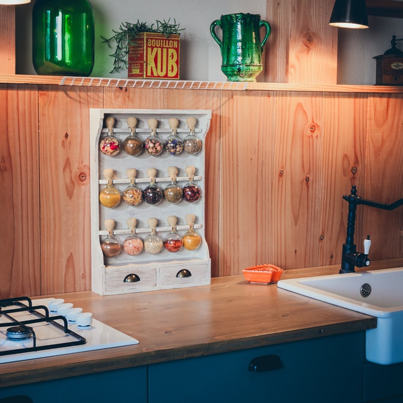 May include: White wooden spice rack with 18 clear glass jars filled with various spices and herbs. The rack has two drawers and is mounted on a wooden wall.
