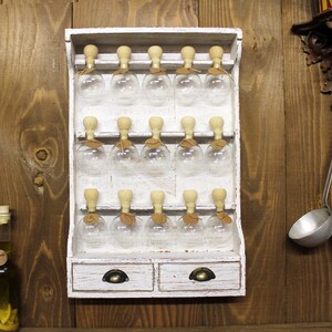 May include: White wooden spice rack with 18 glass jars with cork tops. The rack has two drawers with metal handles.