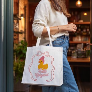 May include: White tote bag with a pink bow and a yellow rubber duck with a pink bow on top. The text "Hello Gorgeous" is printed in pink on the bag.