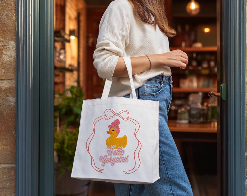 May include: White tote bag with a pink bow and a yellow rubber duck with a pink bow on top. The text "Hello Gorgeous" is printed in pink on the bag.