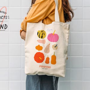 May include: A white canvas tote bag with a colorful illustration of pumpkins and gourds. The text "It's the most... PUMPKIN season of all" is printed on the bag.