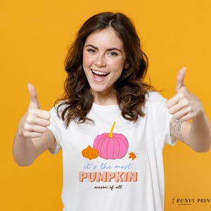 May include: A woman wearing a white t-shirt with a pink and orange pumpkin graphic and the text "it's the most PUMPKIN season of all". She is giving a thumbs up with both hands and smiling.