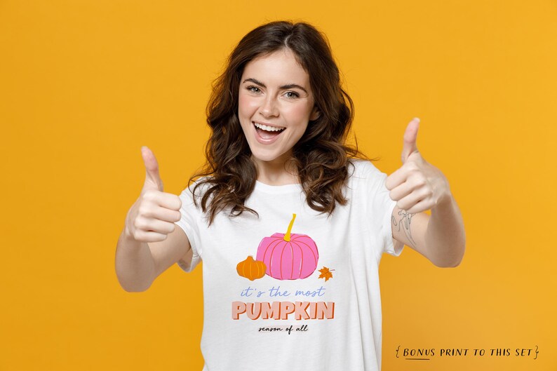 May include: A woman wearing a white t-shirt with a pink and orange pumpkin graphic and the text "it's the most PUMPKIN season of all". She is giving a thumbs up with both hands and smiling.