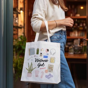 Puede incluir: Una bolsa de tela blanca con una ilustraci&oacute;n colorida de art&iacute;culos de bienestar, que incluyen una planta, un libro, una botella de agua, una esterilla de yoga y unos auriculares. El texto "Wellness Girl" est&aacute; impreso en la bolsa.