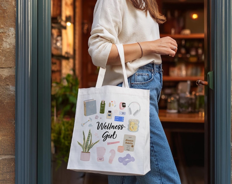Puede incluir: Una bolsa de tela blanca con una ilustraci&oacute;n colorida de art&iacute;culos de bienestar, que incluyen una planta, un libro, una botella de agua, una esterilla de yoga y unos auriculares. El texto "Wellness Girl" est&aacute; impreso en la bolsa.