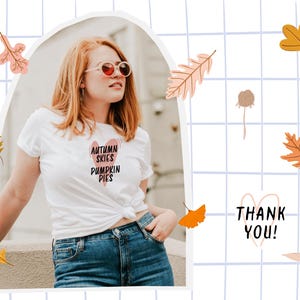 May include: White t-shirt with the text "AUTUMN SKIES PUMPKIN PIES" in a heart-shaped design. The image includes fall-themed illustrations of leaves and a "THANK YOU!" message. The person is wearing blue jeans and sunglasses.