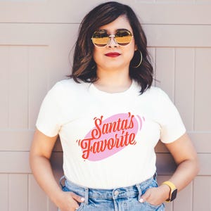 May include: Cream-colored t-shirt with the text "Santa's Favorite" in red script inside a pink oval. The person is wearing gold-rimmed sunglasses, gold hoop earrings, and a yellow watch. The background is a light brown wall.