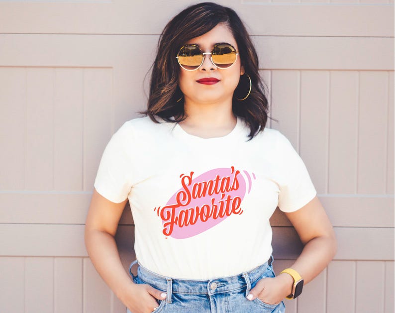 May include: Cream-colored t-shirt with the text "Santa's Favorite" in red script inside a pink oval. The person is wearing gold-rimmed sunglasses, gold hoop earrings, and a yellow watch. The background is a light brown wall.