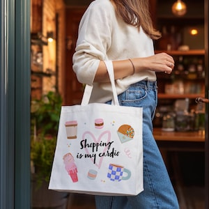 K&ouml;nnte beinhalten: Wei&szlig;e Tragetasche mit bunten Illustrationen von Kaffee, Eis und Geb&auml;ck. Die Tasche tr&auml;gt den Text "Shopping is my cardio" und ein rosa Herzdesign. Die Tasche wird von einer Person getragen, die einen wei&szlig;en Pullover und blaue Jeans tr&auml;gt.