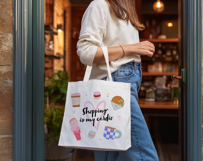 K&ouml;nnte beinhalten: Wei&szlig;e Tragetasche mit bunten Illustrationen von Kaffee, Eis und Geb&auml;ck. Die Tasche tr&auml;gt den Text "Shopping is my cardio" und ein rosa Herzdesign. Die Tasche wird von einer Person getragen, die einen wei&szlig;en Pullover und blaue Jeans tr&auml;gt.