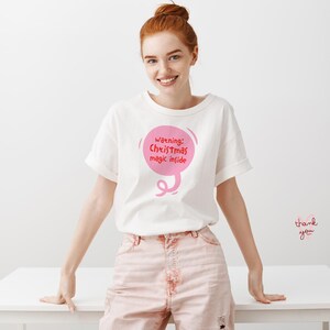 May include: White t-shirt with rolled sleeves featuring a pink speech bubble graphic that reads "Warning: Christmas magic inside." The model is wearing pink pants and standing in front of a white table.