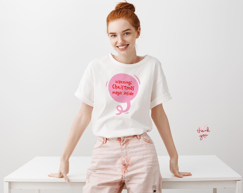May include: White t-shirt with rolled sleeves featuring a pink speech bubble graphic that reads "Warning: Christmas magic inside." The model is wearing pink pants and standing in front of a white table.