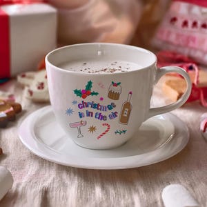 K&ouml;nnte beinhalten: Wei&szlig;e Keramiktasse mit einem festlichen Design mit dem Text "Christmas is in the air" und bunten Illustrationen eines Weihnachtsbaums, eines Strumpfes, eines Zuckerstabs, einer Weinflasche und eines Weihnachtspudding. Die Tasse ist mit einer hellbraunen Fl&uuml;ssigkeit gef&uuml;llt und steht auf einem wei&szlig;en Unterteller.