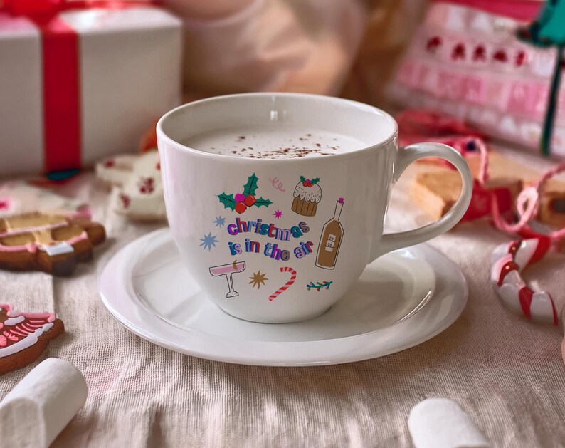 K&ouml;nnte beinhalten: Wei&szlig;e Keramiktasse mit einem festlichen Design mit dem Text "Christmas is in the air" und bunten Illustrationen eines Weihnachtsbaums, eines Strumpfes, eines Zuckerstabs, einer Weinflasche und eines Weihnachtspudding. Die Tasse ist mit einer hellbraunen Fl&uuml;ssigkeit gef&uuml;llt und steht auf einem wei&szlig;en Unterteller.