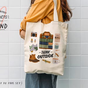 May include: A white canvas tote bag with a graphic design featuring a plaid shirt, boots, a map, a camera, a mug, and the text "Think Outside".