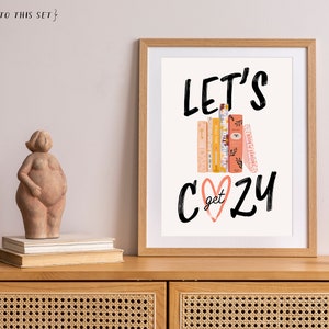 May include: A framed print with the text "Let's get cozy" in black script font. The text is layered over a stack of books with a pink heart in the center. The print is on a white background and has a light wood frame.