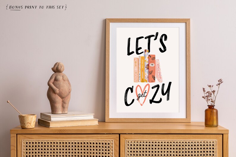 May include: A framed print with the text "Let's get cozy" in black script font. The text is layered over a stack of books with a pink heart in the center. The print is on a white background and has a light wood frame.
