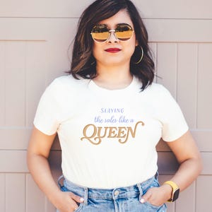 K&ouml;nnte beinhalten: Cremefarbenes T-Shirt mit dem Text "Slaying the sales like a QUEEN" in Gold und Blau. Die Person tr&auml;gt eine blaue Jeans, eine goldfarbene Sonnenbrille und eine gelbe Uhr.