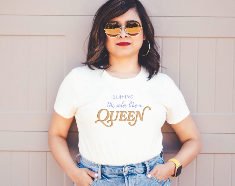 K&ouml;nnte beinhalten: Cremefarbenes T-Shirt mit dem Text "Slaying the sales like a QUEEN" in Gold und Blau. Die Person tr&auml;gt eine blaue Jeans, eine goldfarbene Sonnenbrille und eine gelbe Uhr.