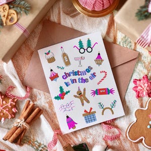 K&ouml;nnte beinhalten: Eine wei&szlig;e Karte mit bunten Illustrationen von weihnachtlichen Motiven, darunter ein Lebkuchenmann, Zuckerstangen und ein Weihnachtsbaum. Der Text "Christmas is in the air" ist in einer bunten Schrift geschrieben.