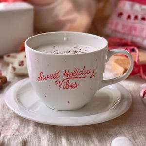 May include: A white ceramic mug filled with a creamy beverage, sprinkled with cocoa, and resting on a matching saucer. The mug features the red text "Sweet Holiday Vibes." Festive cookies, a gift box, and candy canes surround the mug, creating a holiday theme.
