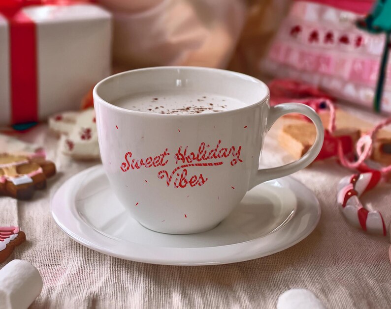 May include: A white ceramic mug filled with a creamy beverage, sprinkled with cocoa, and resting on a matching saucer. The mug features the red text "Sweet Holiday Vibes." Festive cookies, a gift box, and candy canes surround the mug, creating a holiday theme.