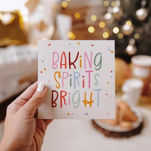 May include: A square card held by a hand, featuring the text "BAKING SPIRITS BRIGHT" in colorful letters. The background is blurred, with Christmas decorations and warm lighting.