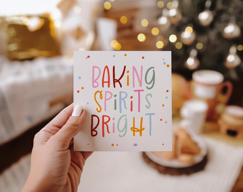 May include: A square card held by a hand, featuring the text "BAKING SPIRITS BRIGHT" in colorful letters. The background is blurred, with Christmas decorations and warm lighting.