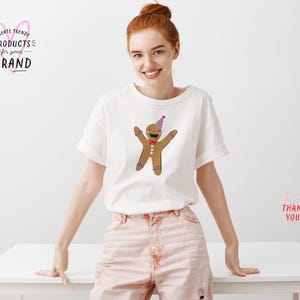 K&ouml;nnte beinhalten: Ein wei&szlig;es T-Shirt mit einem Lebkuchenmann, der einen Partyhut und eine Schleife tr&auml;gt. Der Lebkuchenmann ist braun mit wei&szlig;en Zuckergussdetails. Das T-Shirt wird von einer Frau in rosa Shorts getragen.