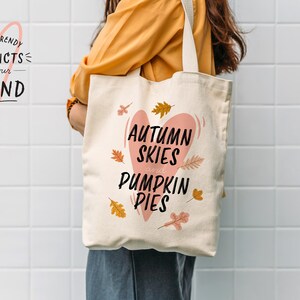 May include: A cream-colored tote bag with the text "Autumn Skies and Pumpkin Pies" in black script inside a pink heart. The bag features fall leaf illustrations and is held by a person wearing a yellow shirt.