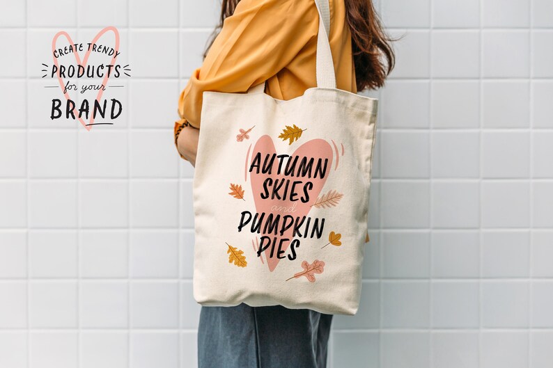 May include: A cream-colored tote bag with the text "Autumn Skies and Pumpkin Pies" in black script inside a pink heart. The bag features fall leaf illustrations and is held by a person wearing a yellow shirt.