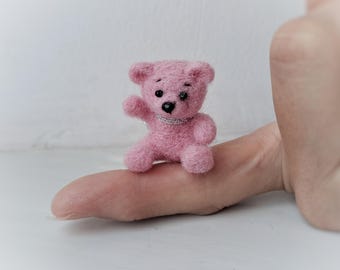 May include: A small pink felt teddy bear with black eyes and a silver necklace. The bear is sitting on a finger.