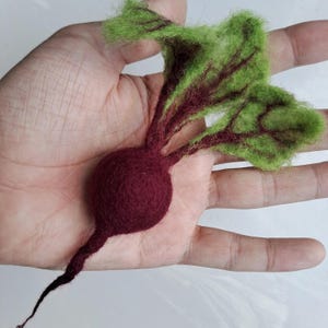 Puede incluir: Una remolacha de fieltro de color rojo intenso con hojas verdes. La remolacha es una representación realista de una remolacha real.