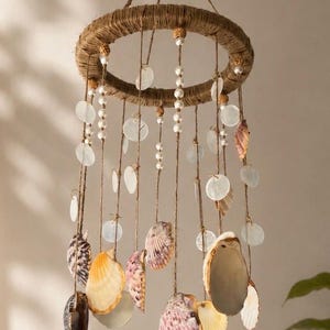 May include: A hanging mobile featuring a circular frame wrapped in brown twine. Multiple strands of twine hang from the ring, decorated with white pearl-like beads, translucent circular shell discs, and various natural seashells in shades of tan, brown, and purple.
