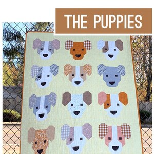 May include: A handmade quilt featuring a repeating pattern of dog faces in various colors and patterns. The quilt is made with patchwork techniques and includes the text "THE PUPPIES" and "A PATTERN BY ELIZABETH HARTMAN".