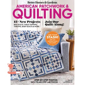 May include: The cover of "American Patchwork & Quilting" magazine features a blue and white quilt draped over a gray sofa. The magazine cover has text that reads "15+ New Projects" and "Join Our Quilt-Along!" A blue pillow and a small side table are also visible.