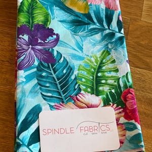 May include: Folded fabric with a tropical floral pattern. The design features large purple orchids, pink hibiscus, and green leaves against a light blue background. A tag with the text "SPINDLE FABRICS" is on top.