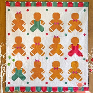 May include: A white quilt with a green and red border. The quilt features 12 gingerbread people in various poses, some wearing bows and some wearing overalls. The gingerbread people are all orange and have white outlines.