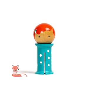 May include: A wooden toy doll with a red head and blue and white polka dot pants. The doll is standing on a blue base.