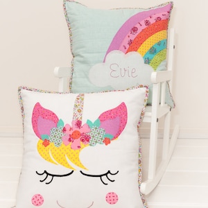 May include: Two decorative pillows, one with a rainbow and a cloud with the name "Evie" embroidered on it, and the other with a unicorn face.