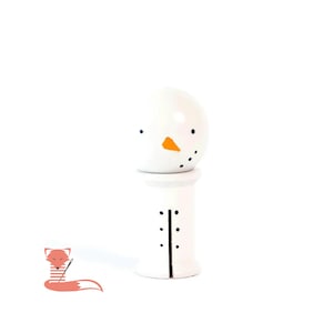 May include: A white snowman-themed decorative object. The top half features a snowman face with black dots for eyes and a mouth, and an orange triangle nose. The bottom half is a white cylinder with black dot and line details.