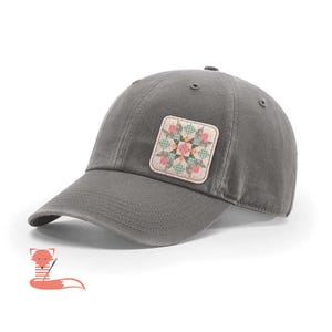 May include: A gray baseball cap with a curved brim. The cap features a square patch with a floral quilt design in pink, green, and peach. The cap is made of a soft material and has a classic, casual style.