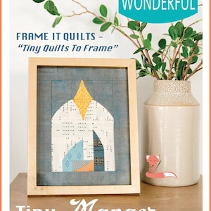 May include: A framed quilt pattern titled "Tiny Manger" with a geometric design in shades of blue, yellow, and orange. The quilt is framed in a gold frame and is sitting on a wooden table. The image also includes a vase with a green plant and a small fox illustration.